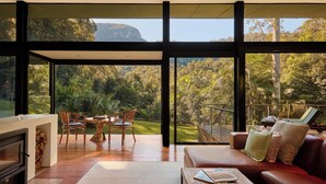 A spacious living area with a fireplace, a dining table with chairs, a sofa, and a large window offering a view of a lush garden and mountains. - Crystal Creek Rainforest Retreat (Upper Crystal Creek)