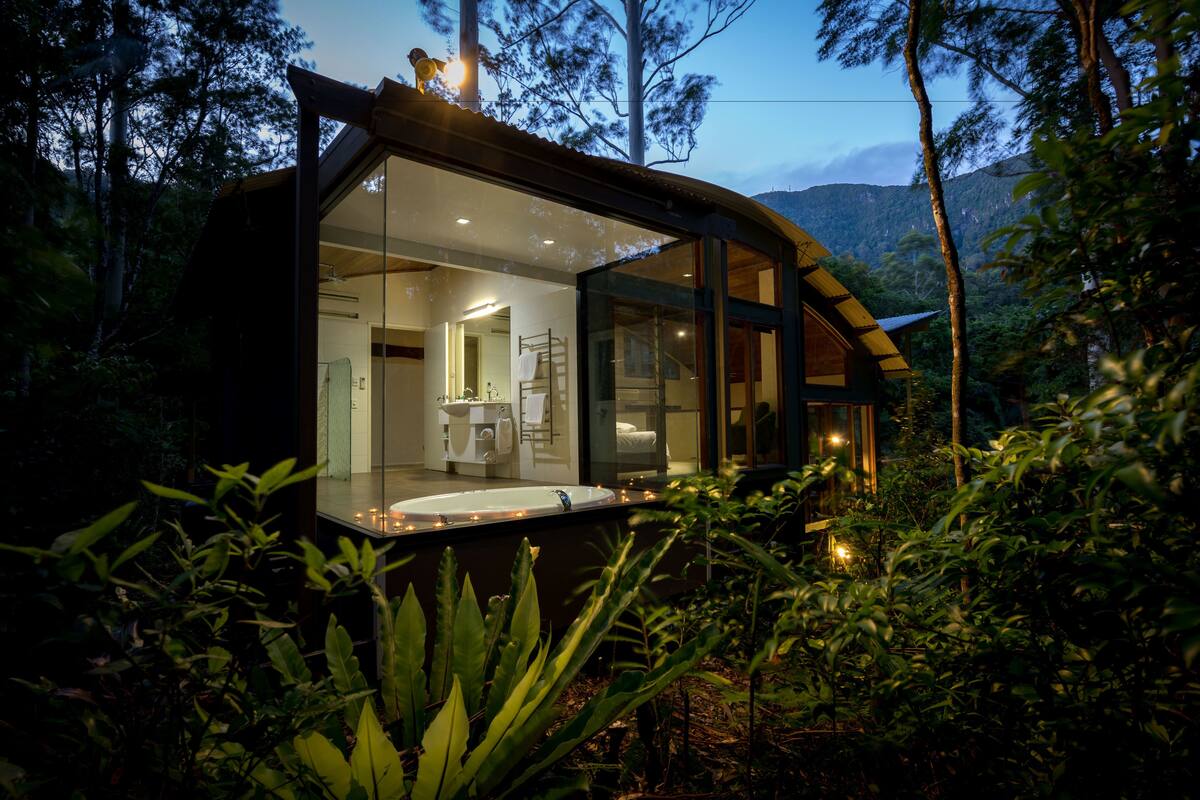 Une cabane moderne, entièrement vitrée, avec une salle de bain visible et une vue sur la forêt environnante.
