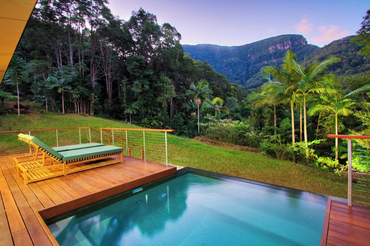 Une terrasse en bois avec une piscine, entourée d’une végétation luxuriante et dominée par un paysage montagneux.