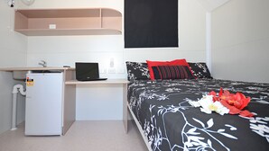 Ein kleines Hotelzimmer mit Bett, Schreibtisch mit Laptop, kleinem Kühlschrank und Fenster mit Verdunkelung.
