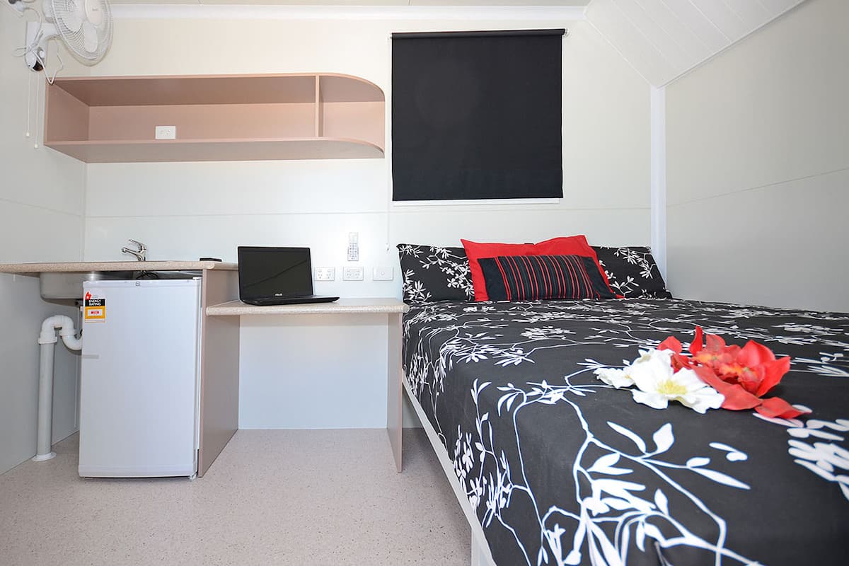 A compact hotel room with a bed, a desk with a laptop, a small refrigerator, and a window with a black blind.