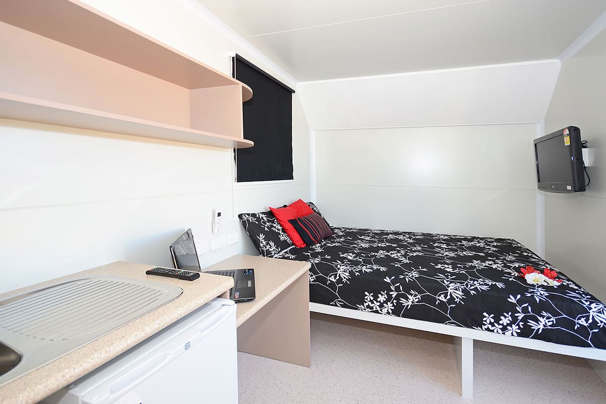 A compact bedroom with a bed, a desk, a small kitchenette, and a TV mounted on the wall.