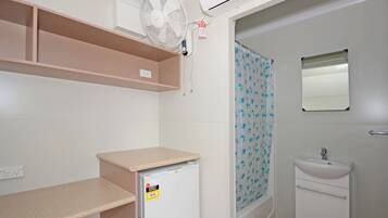 A small, compact bathroom with a white sink, a mirror, a shower with a floral curtain, a small refrigerator, and a ceiling fan.