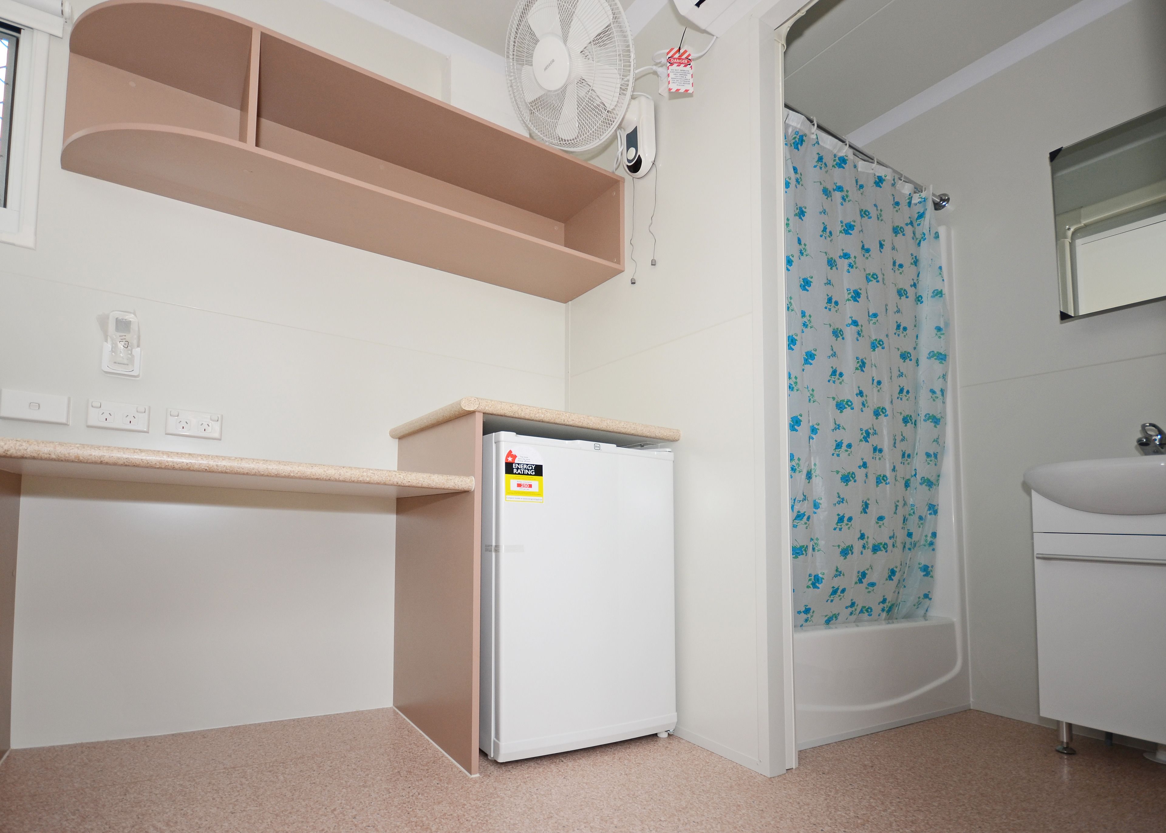 A compact kitchen with a white refrigerator, a small countertop, a fan, and a shower with a floral curtain.