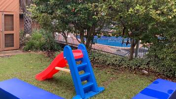 Un parque infantil con un tobogán rojo y una estructura de escalada azul, rodeado de césped y árboles.