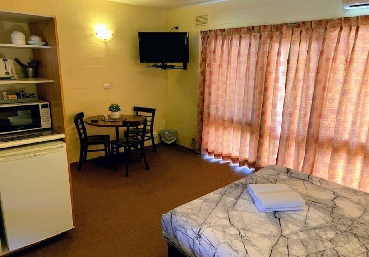 A small, compact room with a bed, a dining table with chairs, a microwave, a refrigerator, and a television mounted on the wall.