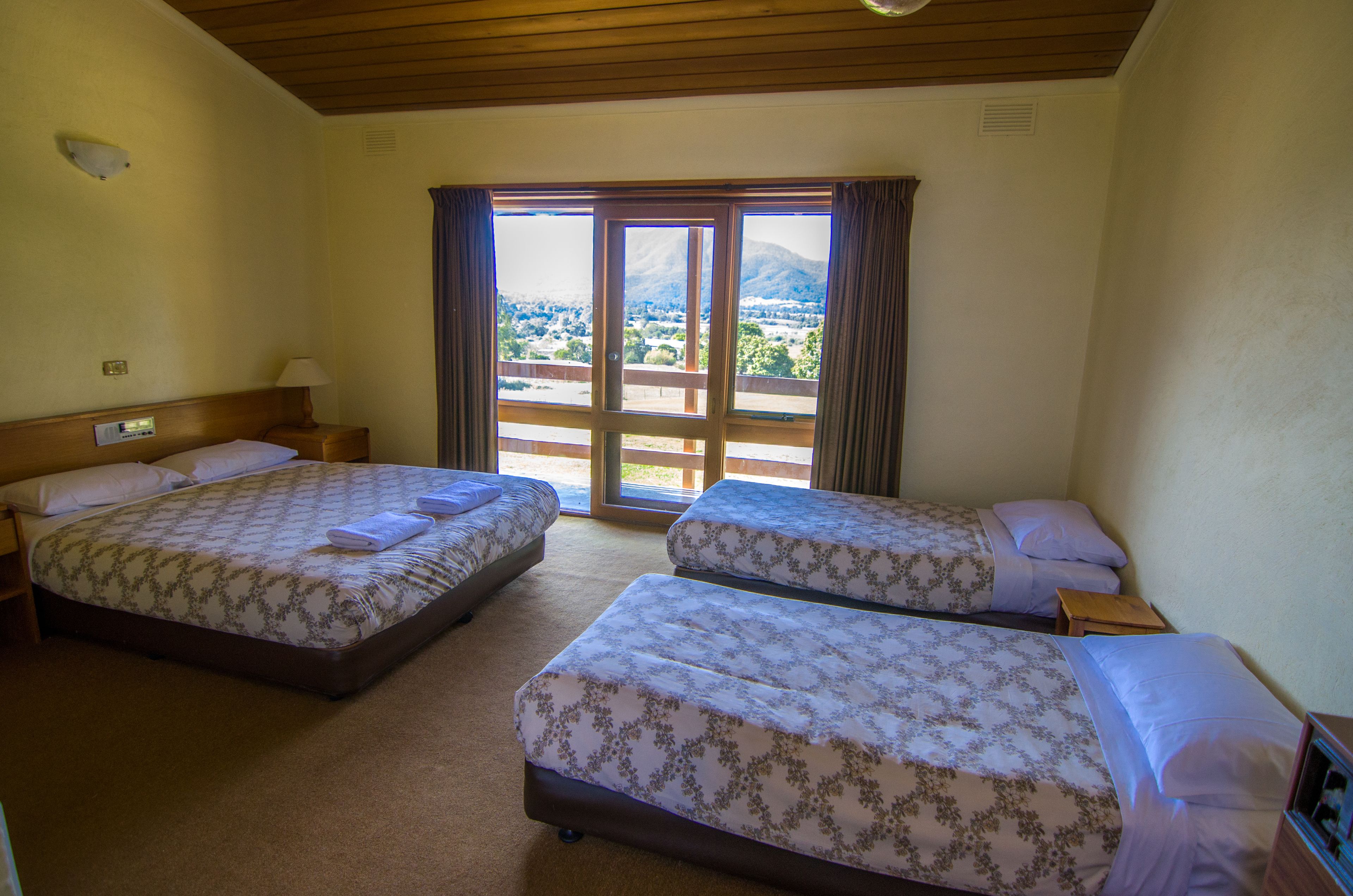 A hotel room with two beds, a wooden ceiling, a view through a large window, and a small table with a lamp.