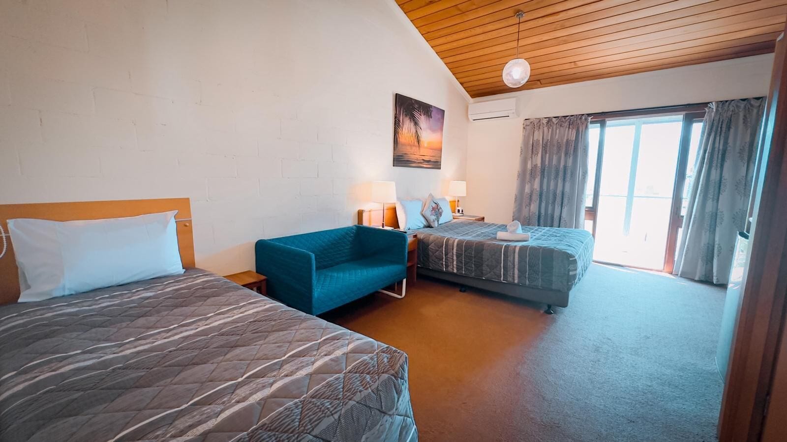 A hotel room with two beds, a blue armchair, a painting on the wall, and a large window with curtains.