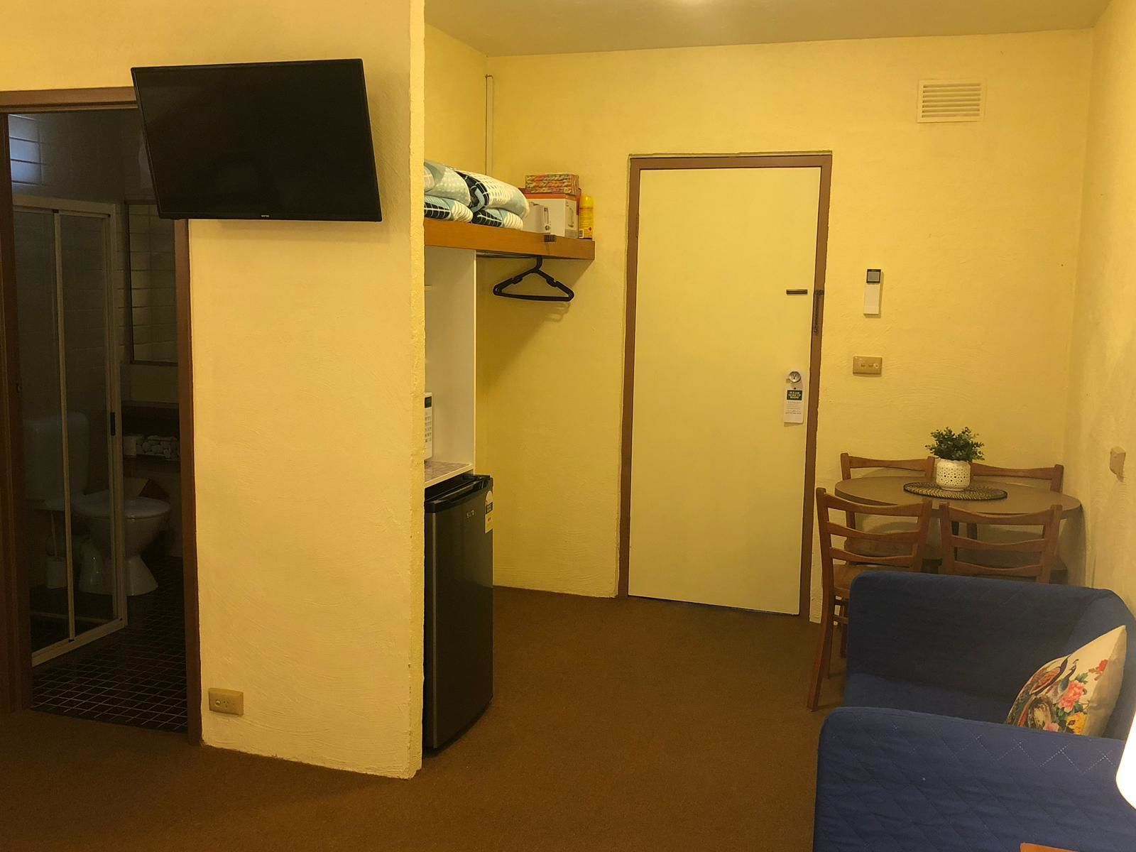 A compact living space with a kitchenette, a television mounted on the wall, a small dining area with a table and chairs, and a door leading to another room.