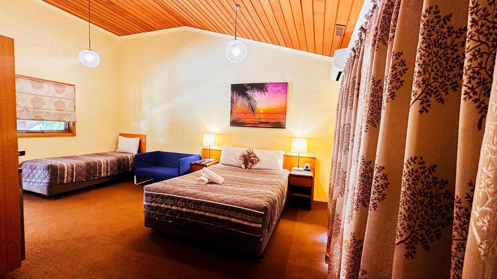 A hotel room with a large bed, a smaller bed, a chair, and a painting of a sunset.