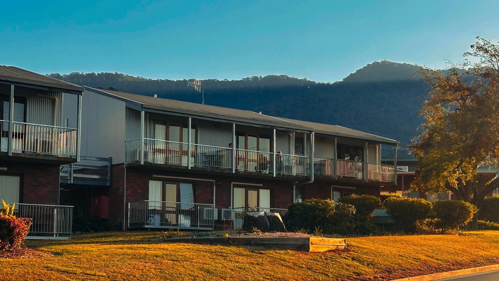 A multi-story motel with balconies, a parking area, and a mountainous backdrop.