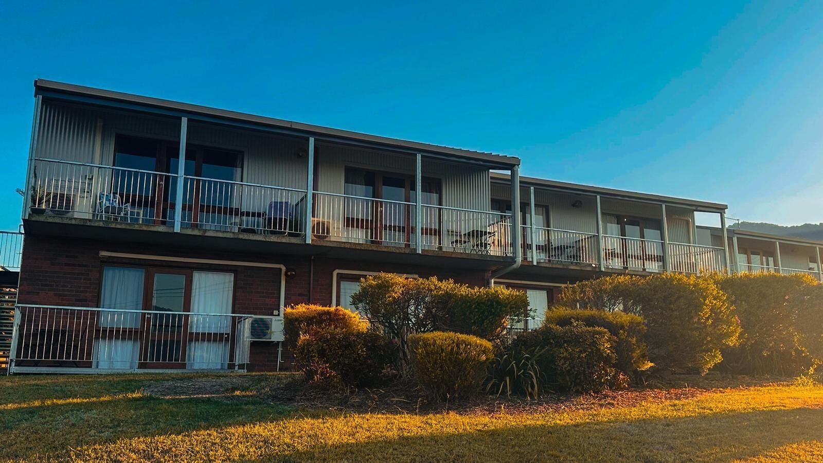 A two-story building with balconies, a brick facade, and a grassy area with bushes.