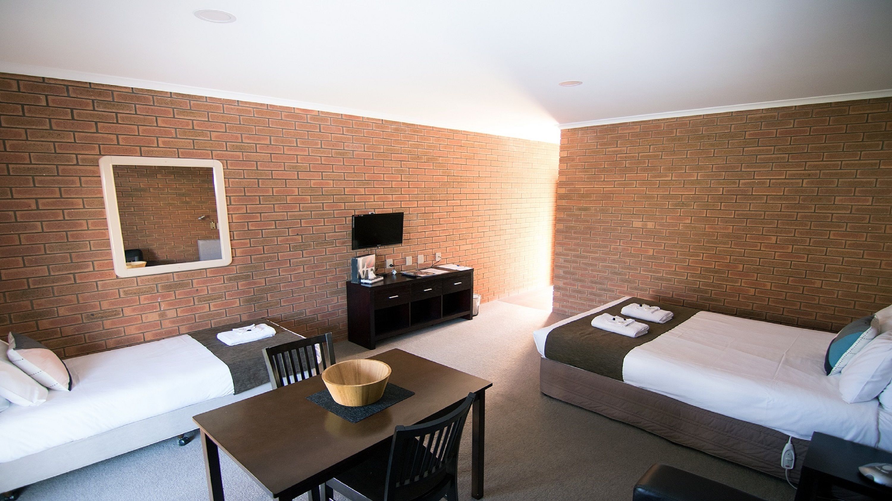 Een hotelkamer met twee eenpersoonsbedden, een klein tafeltje met een schaal, een televisie op een standaard en een bakstenen muur.