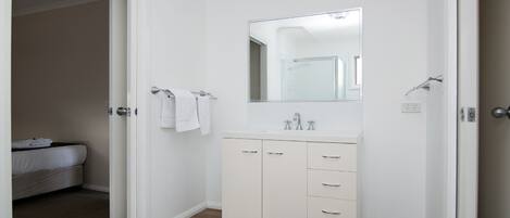 A small, clean bathroom with a white vanity, a mirror, and a bed in the adjacent room.