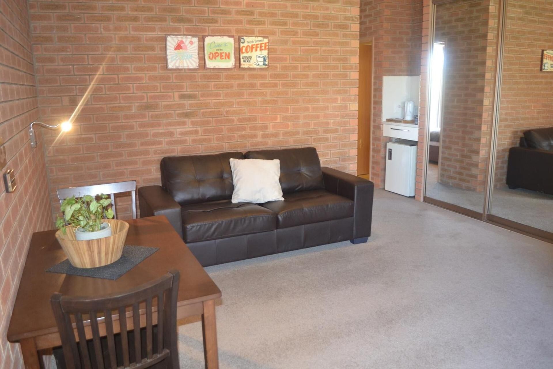 A cozy living room with a brick wall, a brown leather sofa, a wooden table with a plant, and a small white refrigerator.