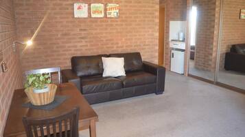 Un salón acogedor con pared de ladrillo, un sofá de cuero marrón, una mesa de madera con una planta y un refrigerador blanco pequeño.