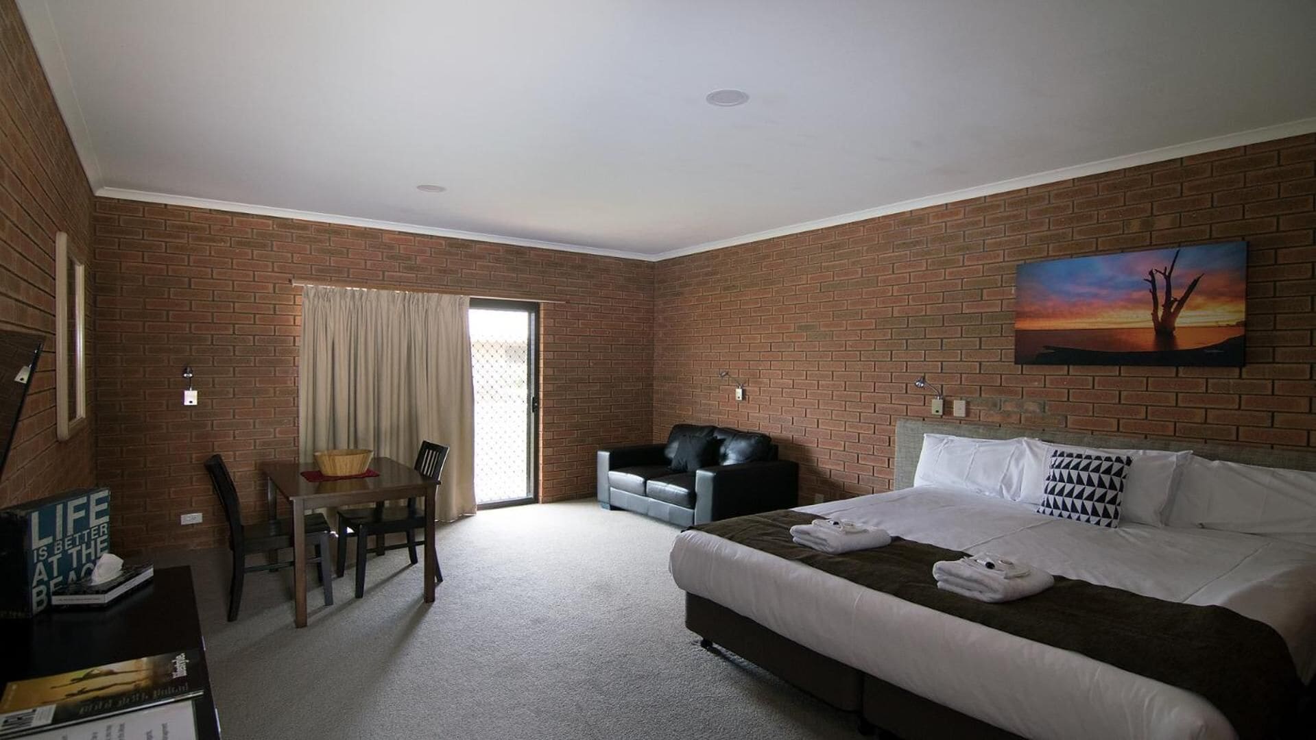 A hotel room with a brick wall, a bed with white bedding, a black leather sofa, a dining table with chairs, and a painting on the wall.