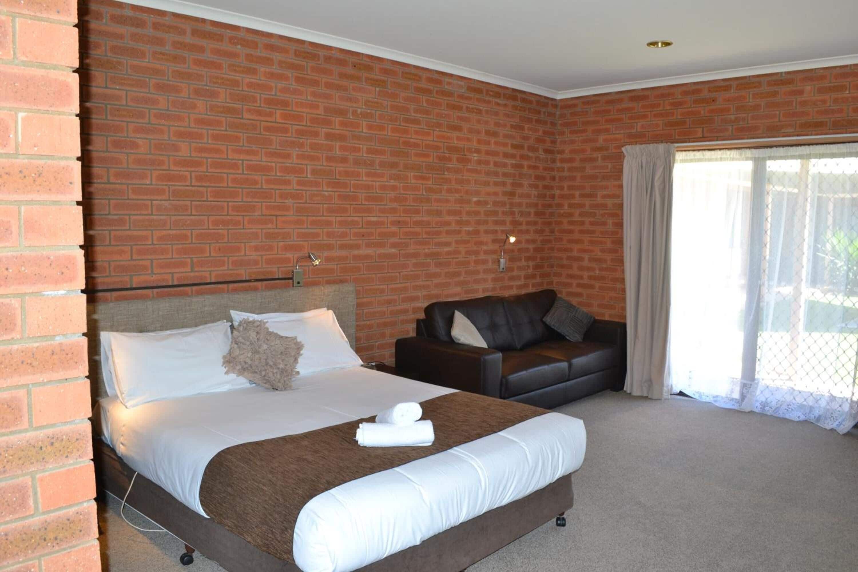 Habitación de hotel con pared de ladrillo, cama con sábanas blancas, sofá de cuero negro y una ventana grande con cortinas.
