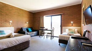 Habitación de hotel con pared de ladrillo, cama con ropa blanca, sofá con almohada azul, escritorio con silla y televisor montado en la pared.