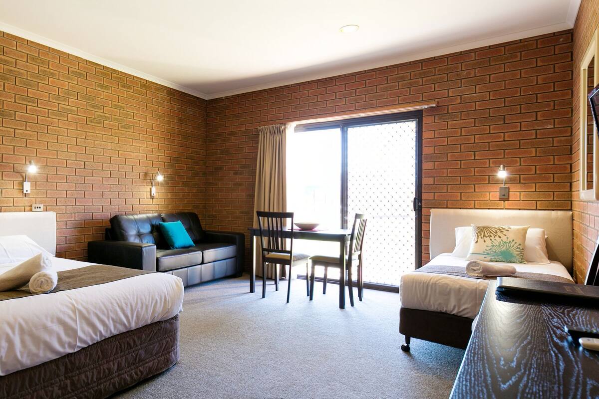 Habitación de hotel con pared de ladrillo, cama con ropa blanca, sofá con almohada azul, escritorio con silla y televisor montado en la pared.