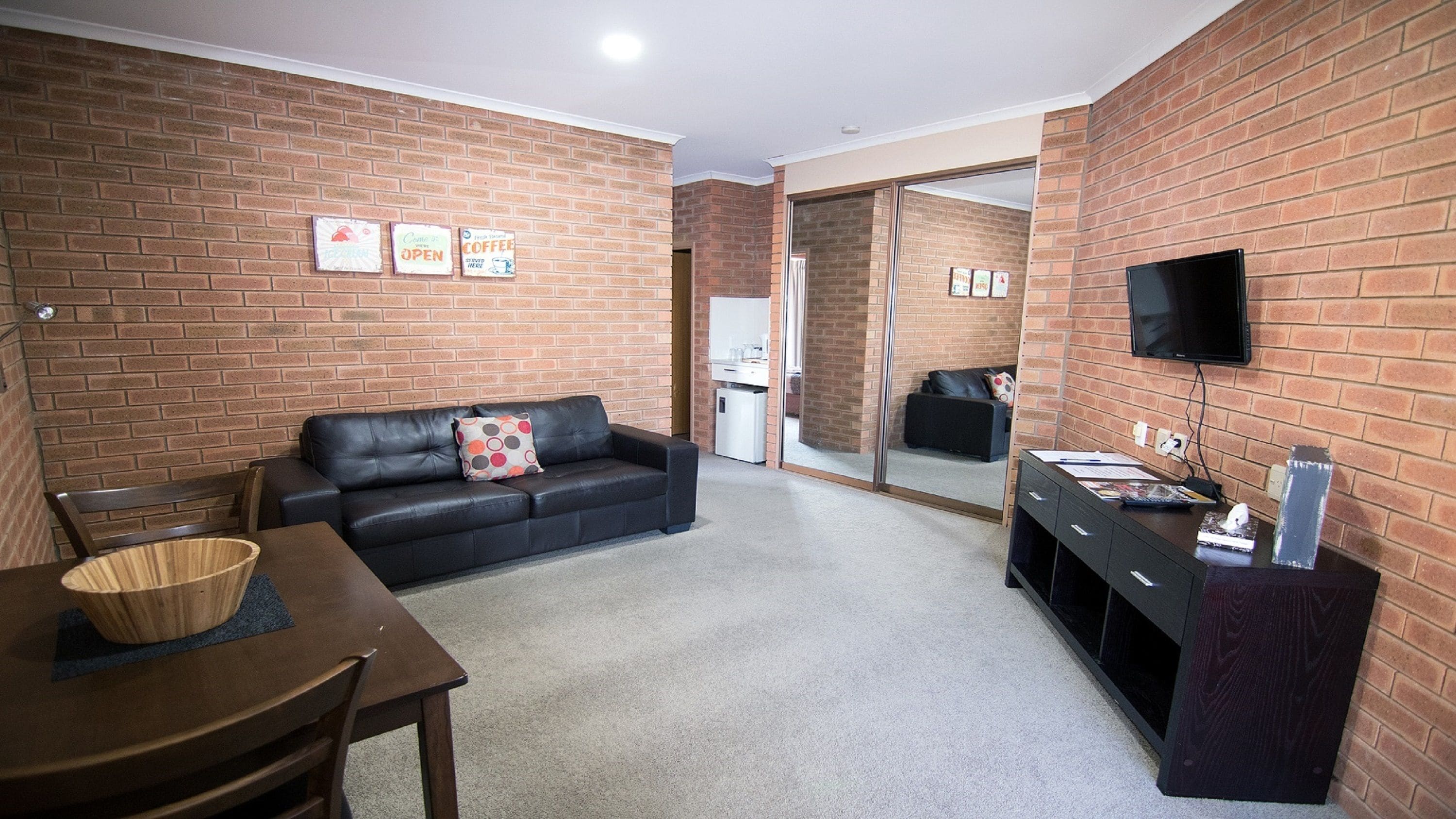 A compact living space with a brick wall, a black leather sofa, a dining table with chairs, a small kitchenette, and a flat-screen TV mounted on the wall.