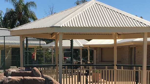 A clear pool with a rock formation in the foreground, a covered seating area with a white roof, and a fenced enclosure in the background. - The Jolly Swagman Motor Inn (Goondiwindi)