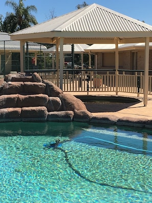 Uma piscina transparente com uma formação rochosa em primeiro plano, uma área de estar coberta com um teto branco e um cercado ao fundo.