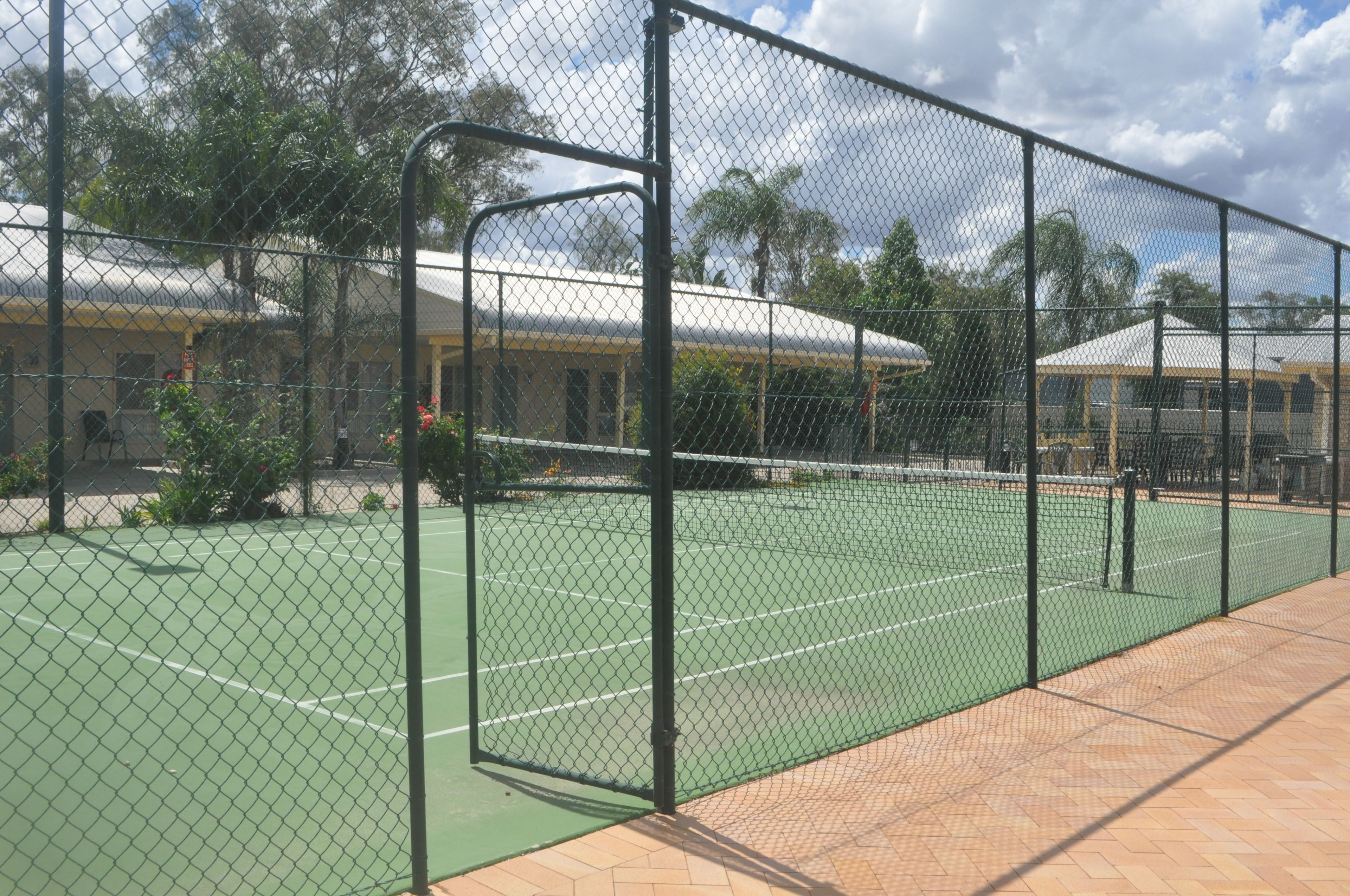 Un court de tennis entouré d’une clôture en treillis métallique, avec un chemin piétonnier en briques.