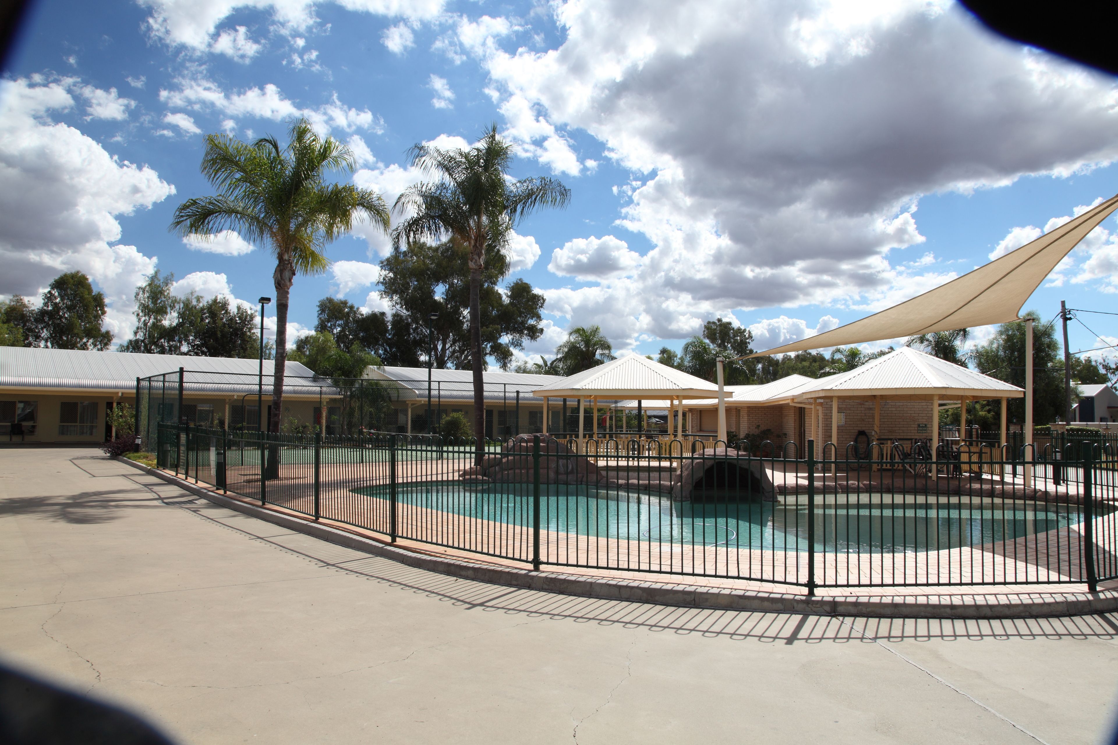 A swimming pool with a fence, palm trees, and covered seating areas.