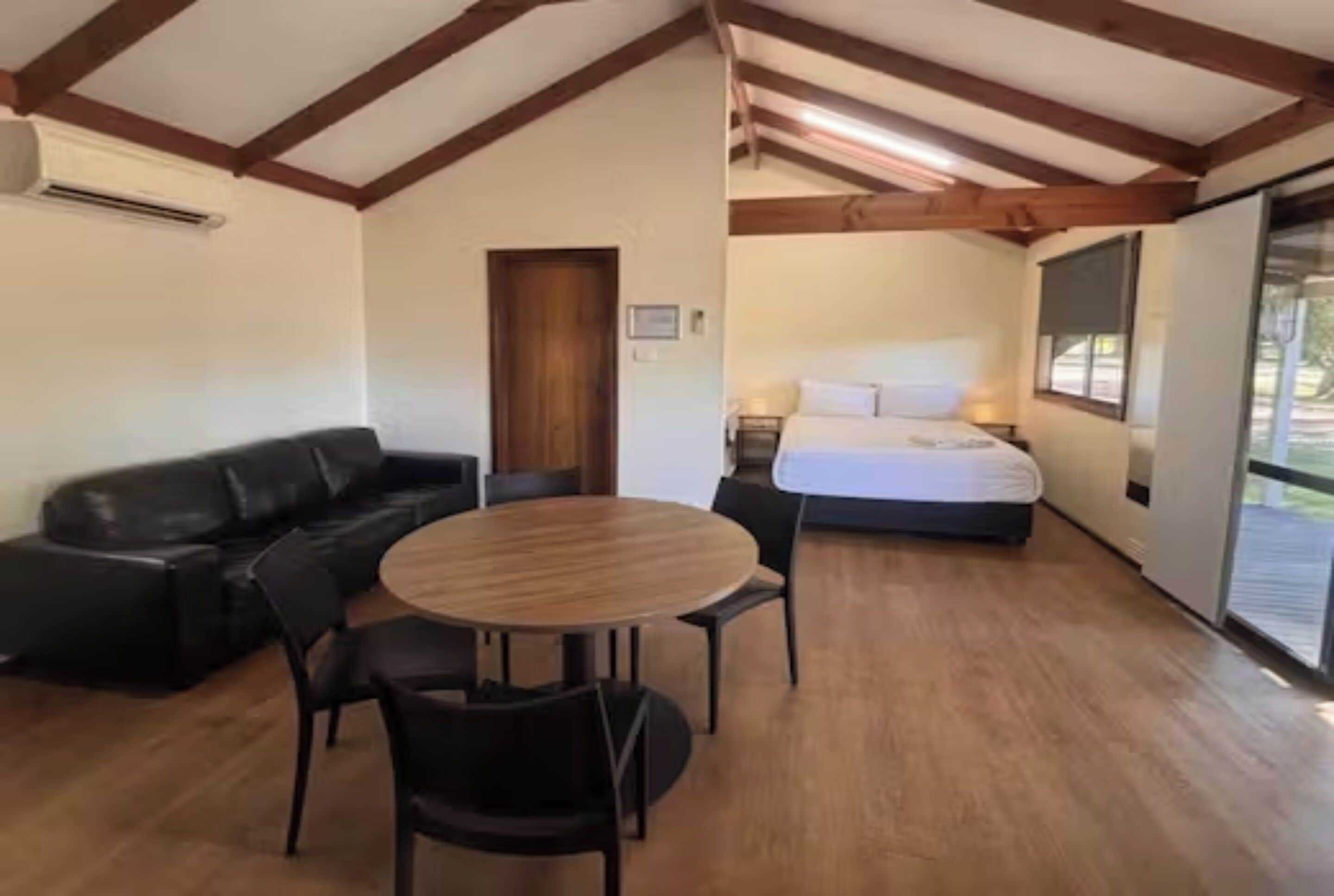 A room with a wooden floor, a round table with chairs, a black leather sofa, a bed with white bedding, and a door.