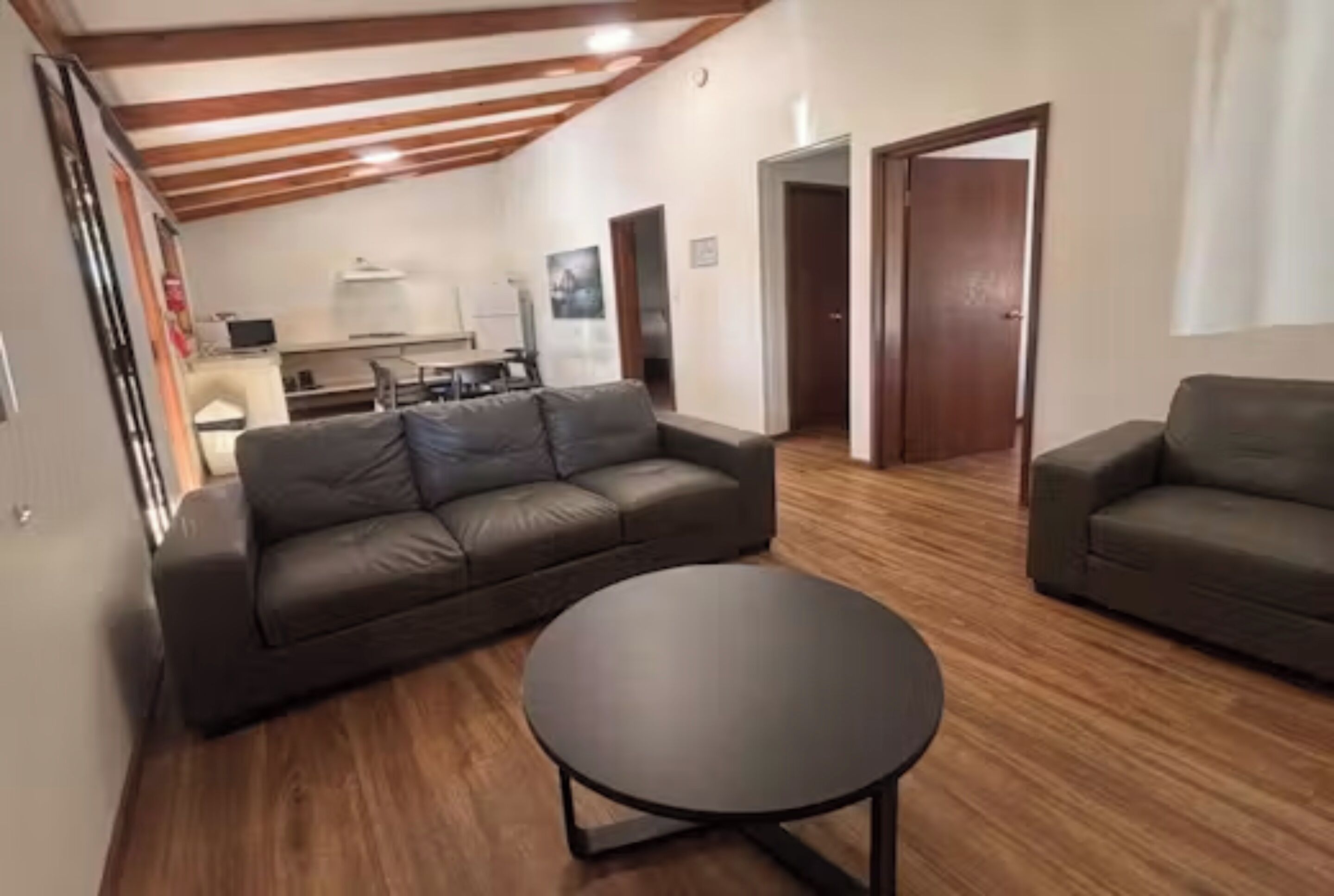 Un salon avec un canapé en cuir, une table basse ronde et un parquet. Au fond, on aperçoit un coin cuisine avec une table à manger et des chaises.