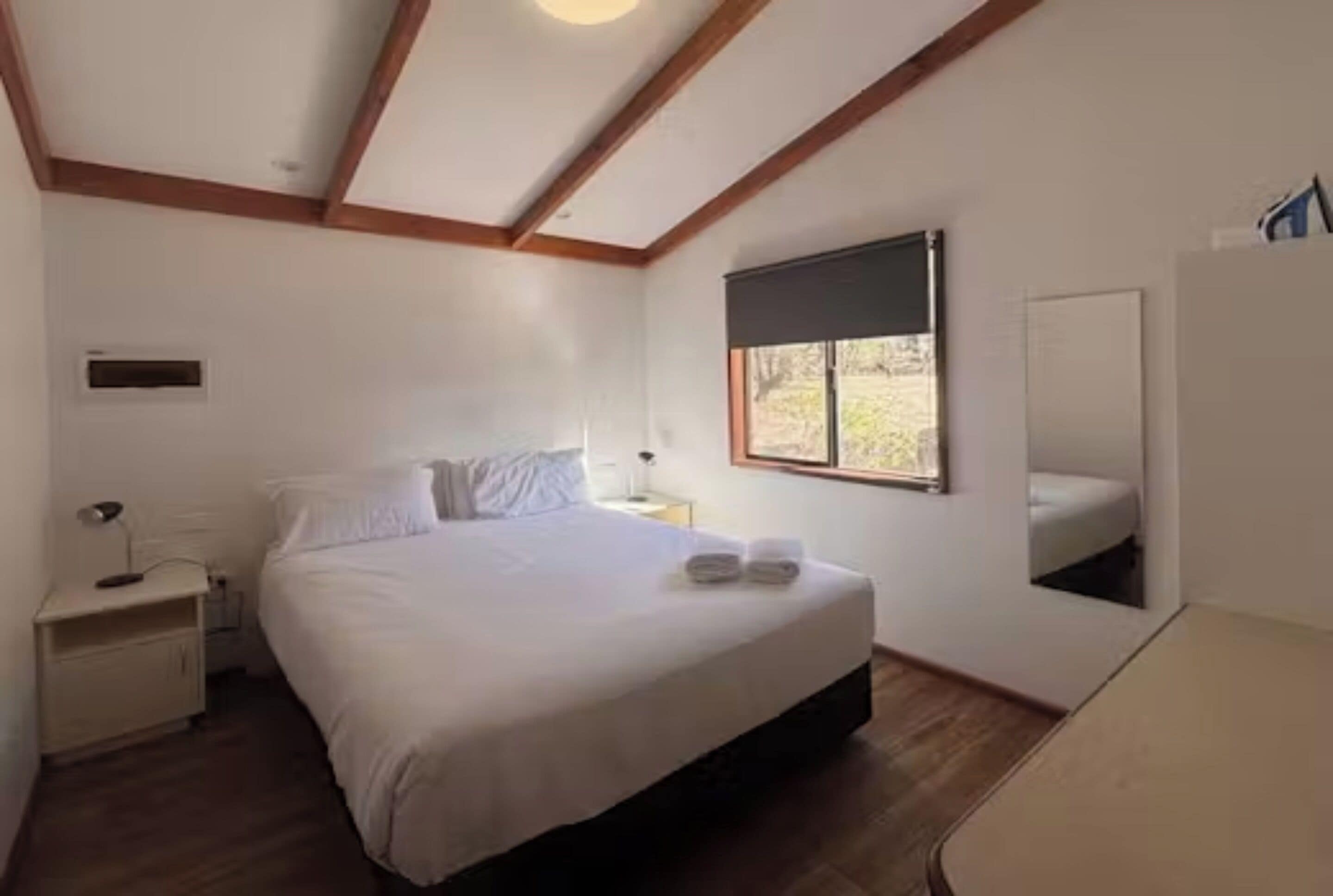Quarto com cama, mesa de cabeceira, abajur, janela e espelho.