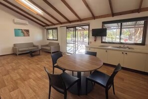 Una habitación con una mesa redonda de madera rodeada de sillas, un sofá, un televisor y una ventana con vista a árboles.