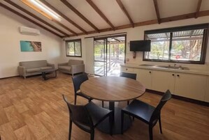 Una habitación con una mesa redonda de madera rodeada de sillas, un sofá, un televisor y una ventana con vista a árboles.