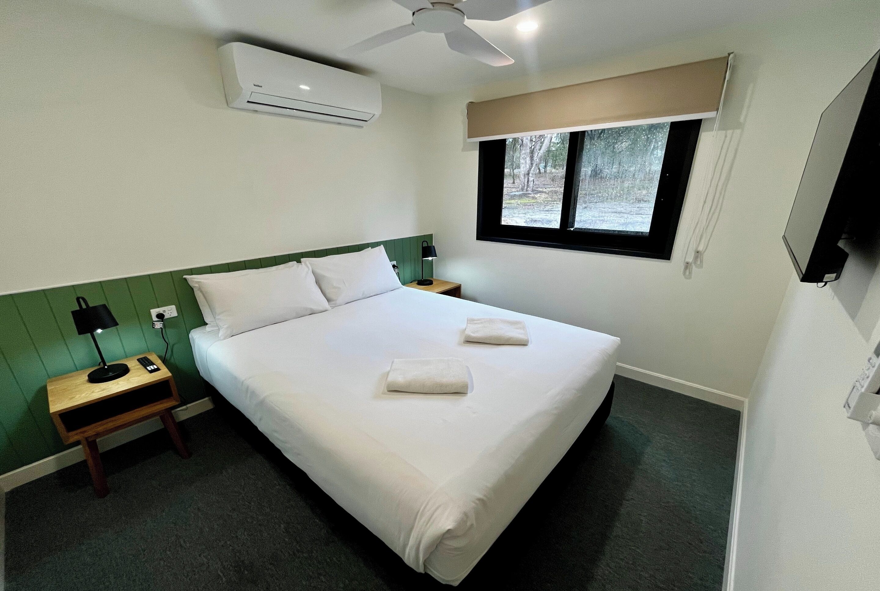 A hotel room with a bed, a nightstand with a lamp, an air conditioner, a window with a view, and a ceiling fan.