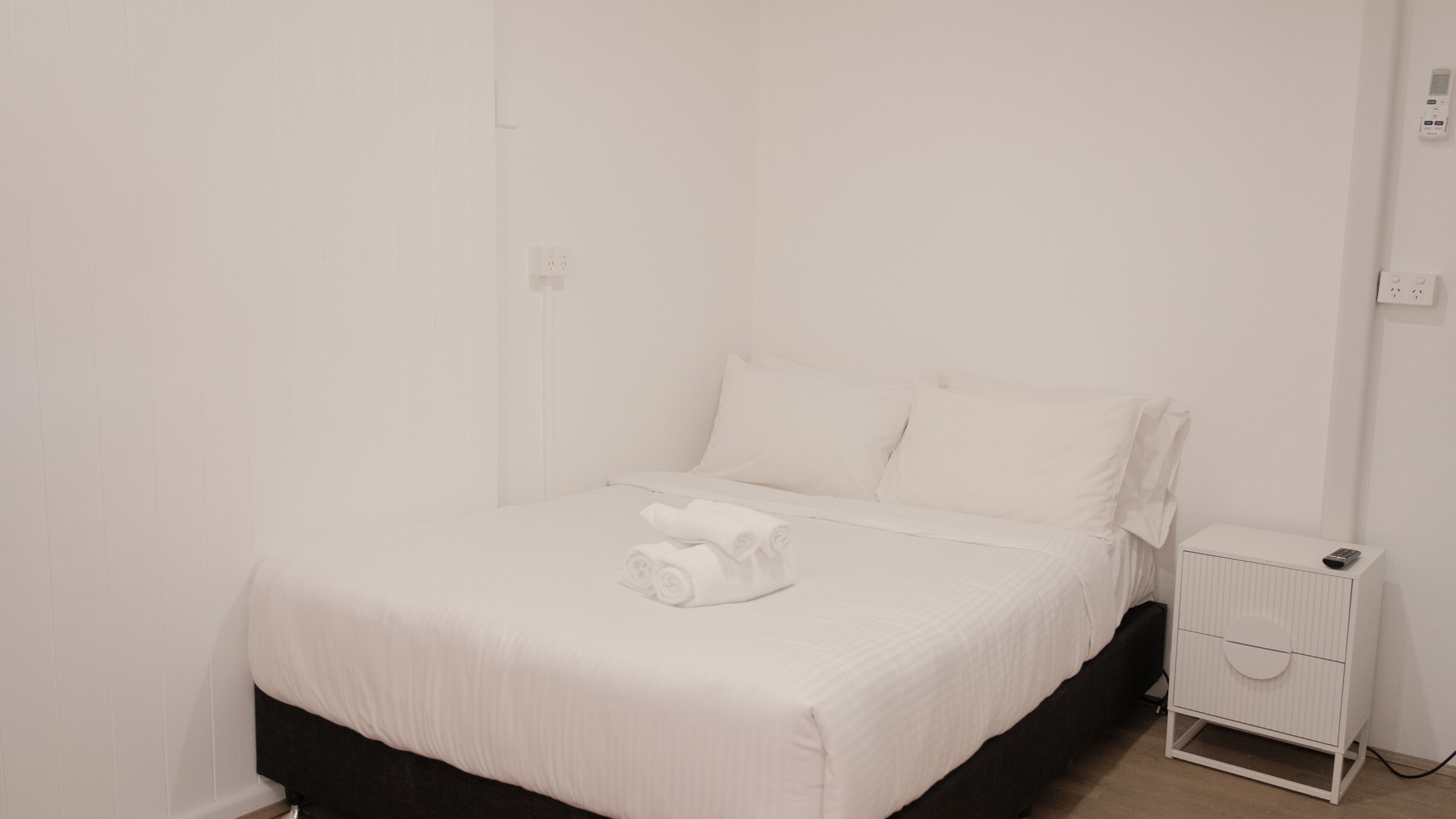 Una habitación blanca con una cama, una mesita de noche y un mueble blanco pequeño.