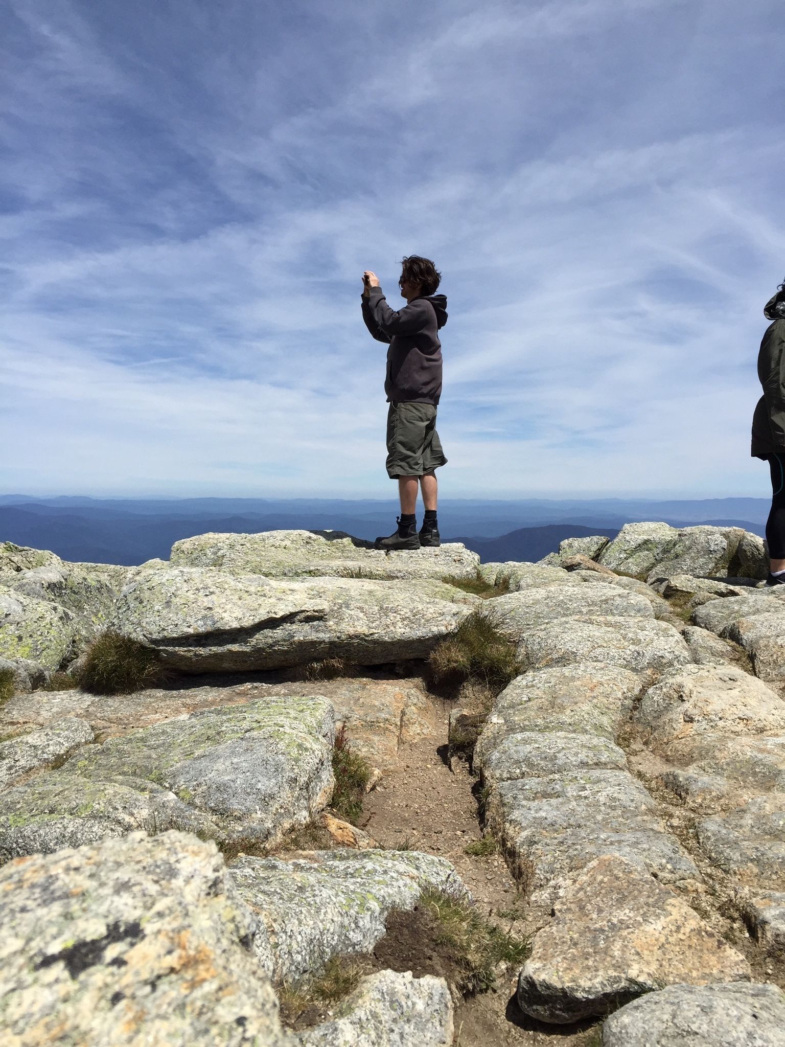 A rocky terrain with two individuals standing on a high vantage point, overlooking a vast landscape.