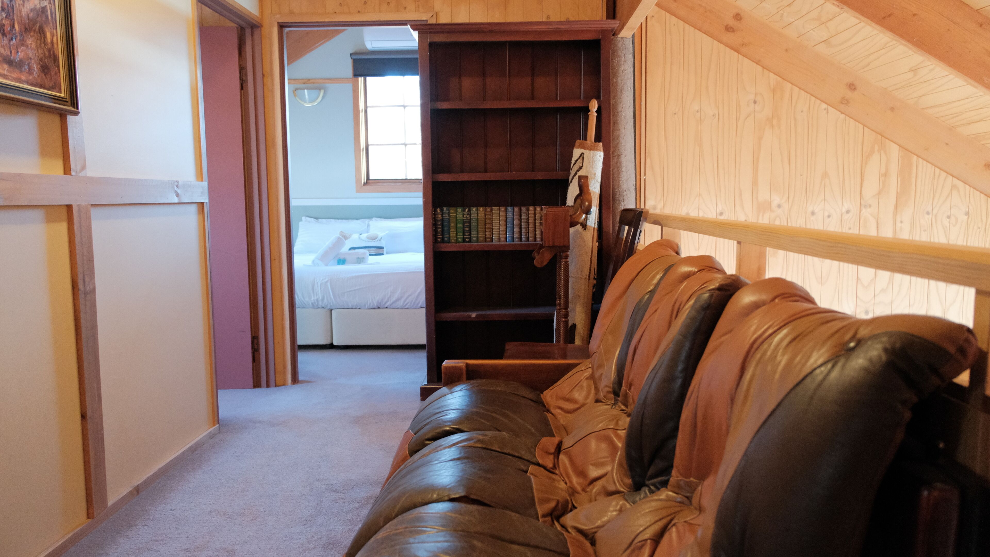 A corridor with a leather sofa, wooden bookshelves, a bed, and a wooden ceiling.