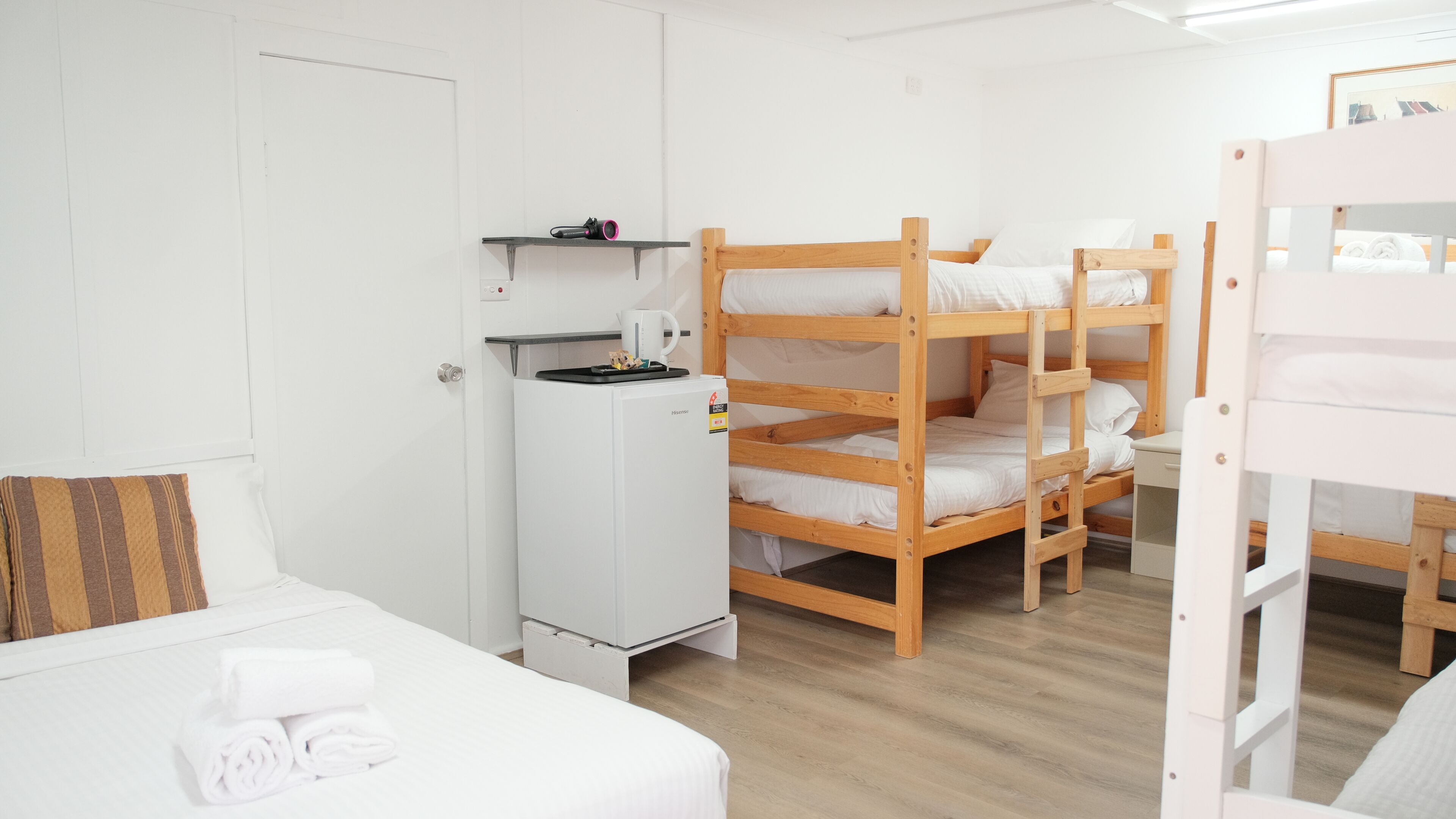 Ein Schlafraum mit Etagenbetten, einem weißen Kühlschrank, einem Bett mit gestreiftem Kopfkissen und Holzboden.