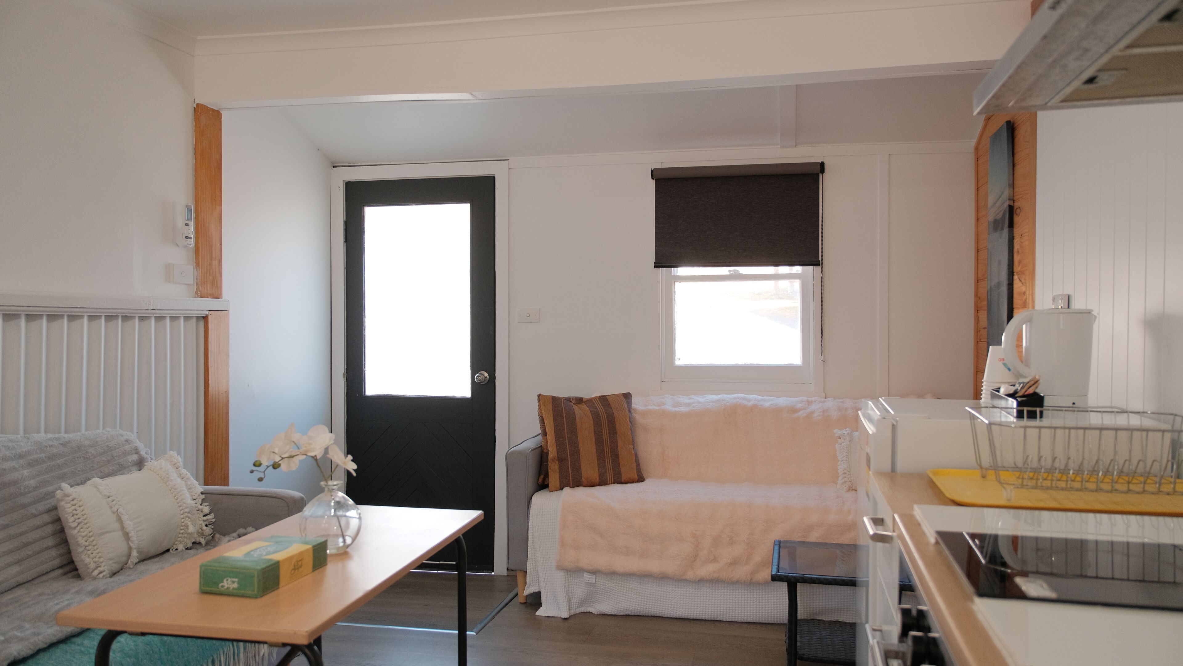 A compact living space with a sofa, a small table with a box, a radiator, a door, a window with a blind, and a kitchenette area.