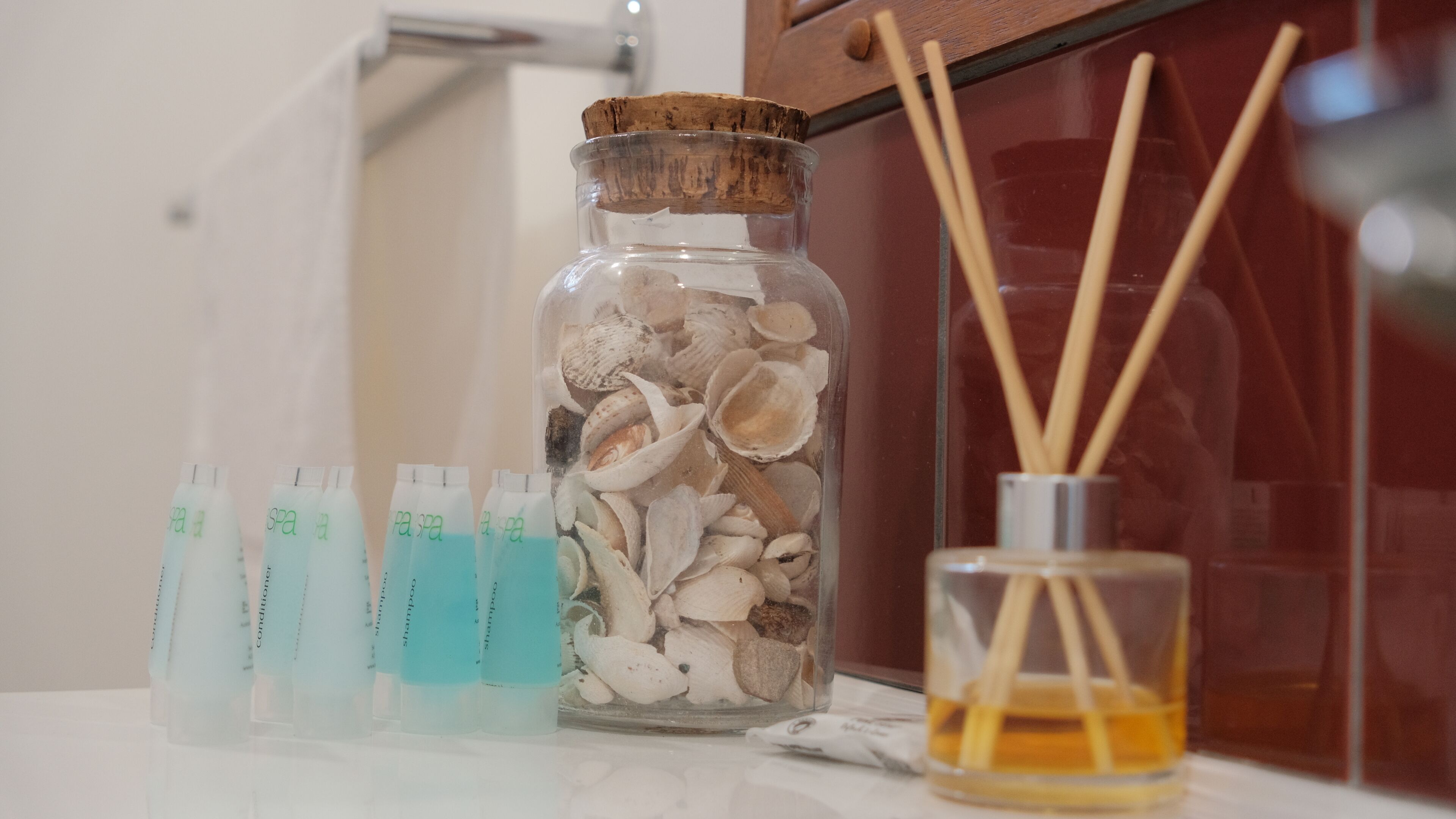 A bathroom with a glass jar filled with seashells, a row of shampoo bottles, and a reed diffuser.