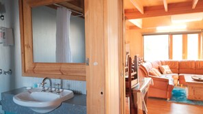Une salle de bain avec un lavabo et un miroir, une porte en bois et une vue sur un espace de vie comprenant un canapé et une table.