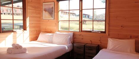 A wooden cabin room with two beds, a nightstand, and a window with a view of a building and landscape.