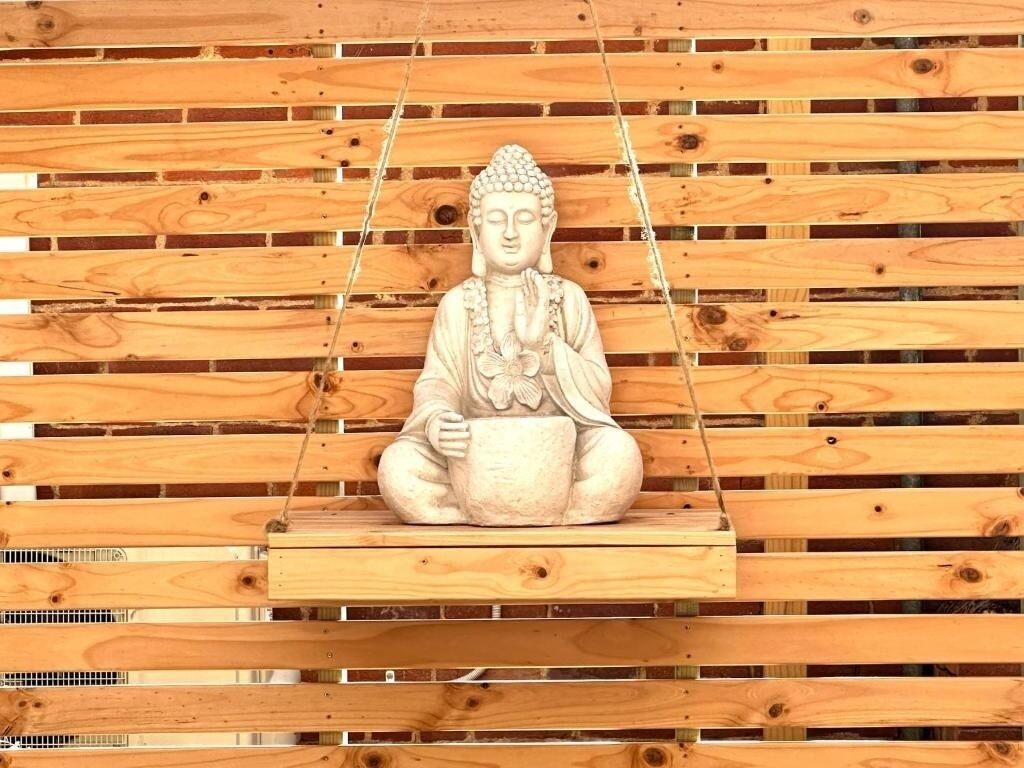 A statue of a seated figure with a bowl, suspended on a wooden platform against a backdrop of horizontal wooden planks.