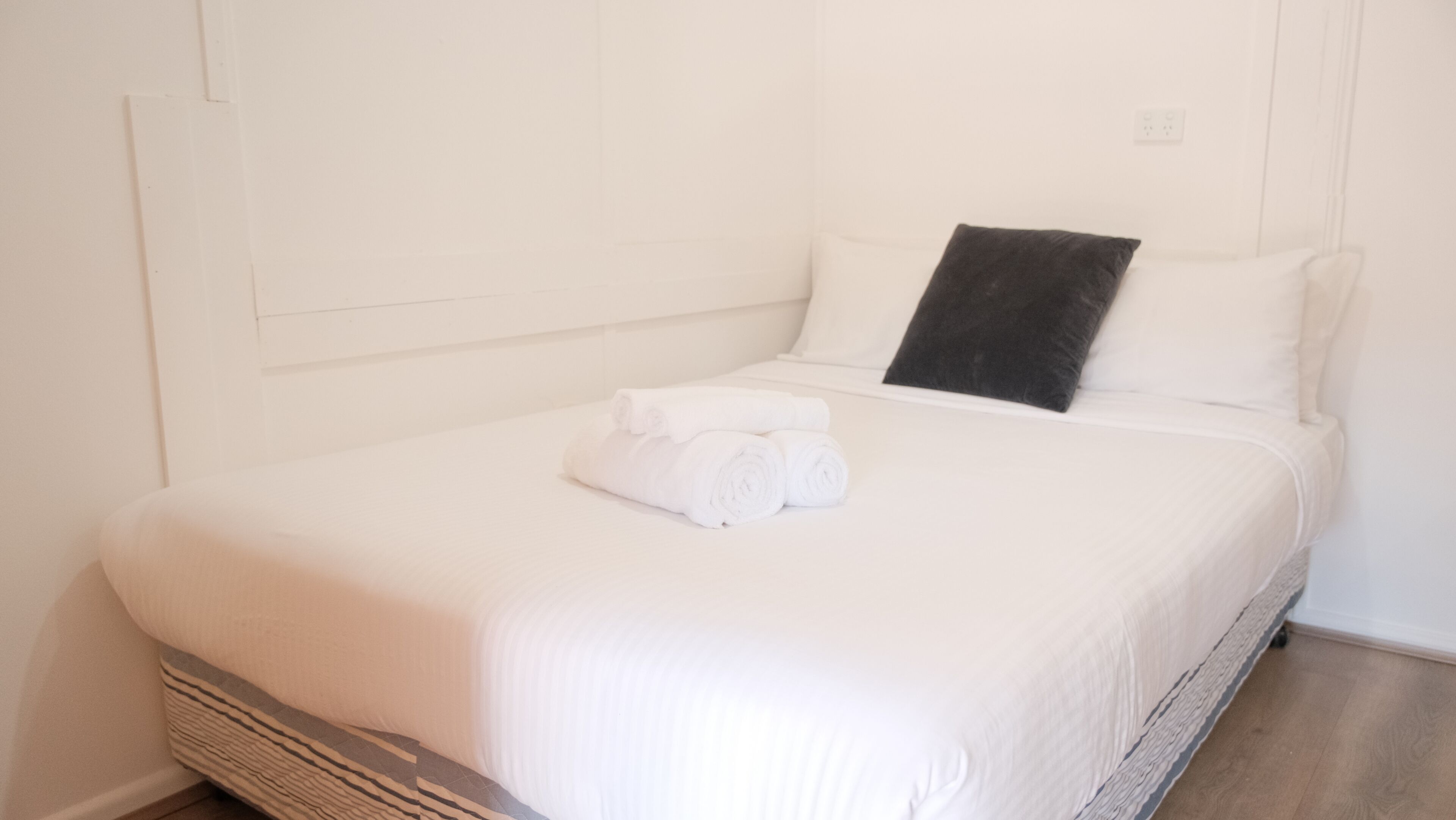 A neatly made bed with white linens and a black pillow, a wooden headboard, and a white wall with a power outlet.