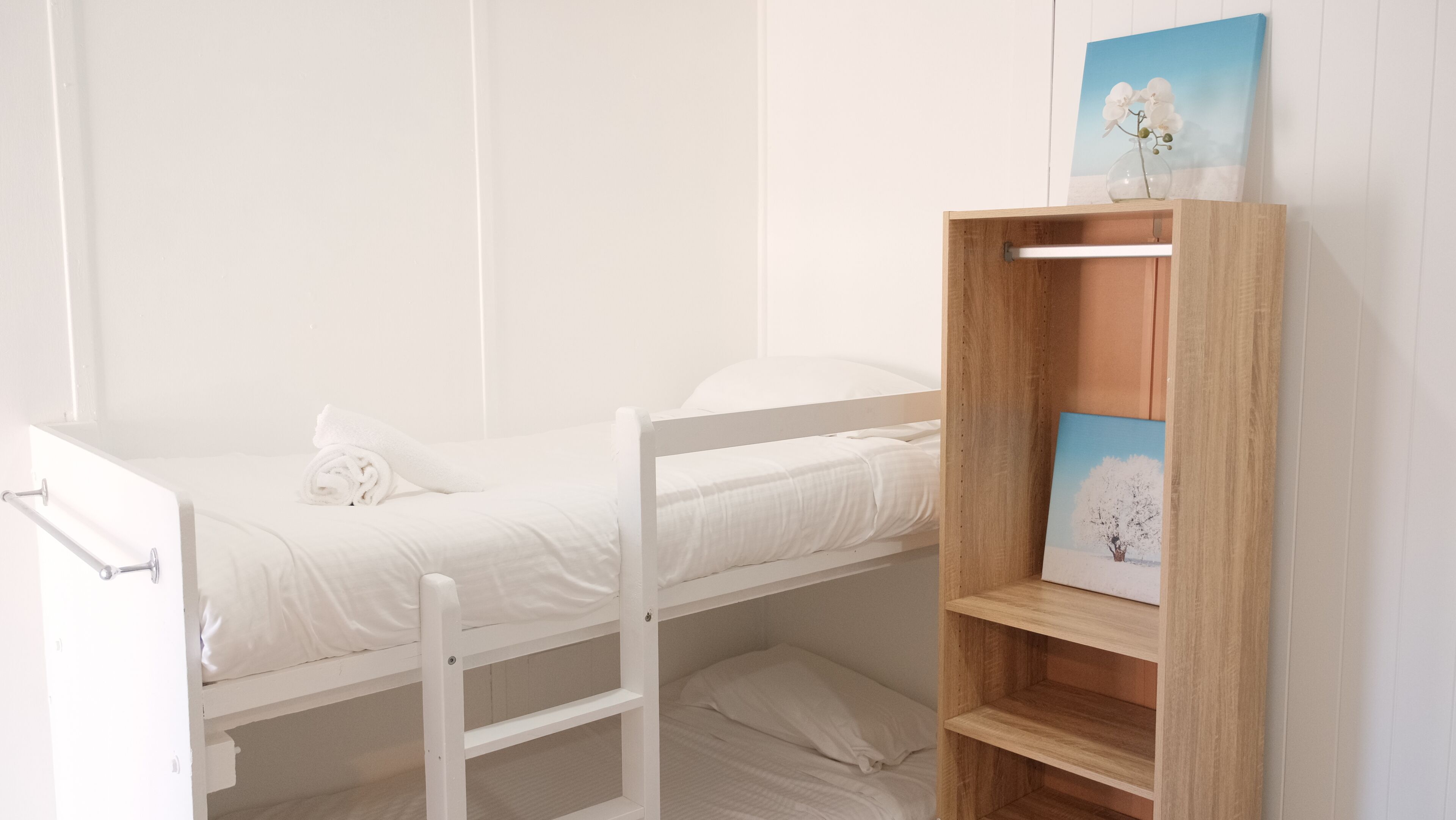 Ein Etagenbett mit weißer Matratze und Kissen, ein Holzregal mit einem Bilderrahmen und eine weiße Tür mit einem Handtuch.