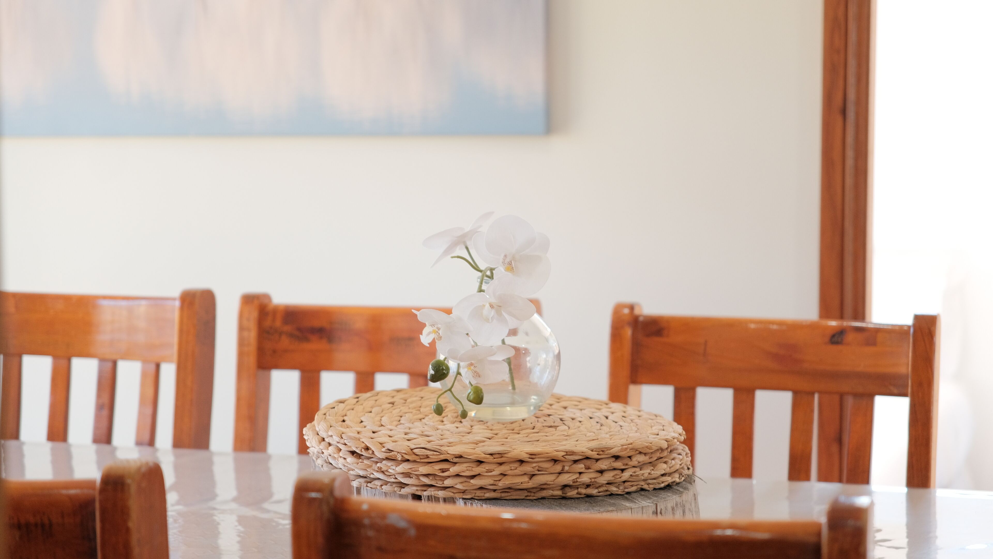 A dining table with a woven placemat, wooden chairs, and a vase with white flowers.