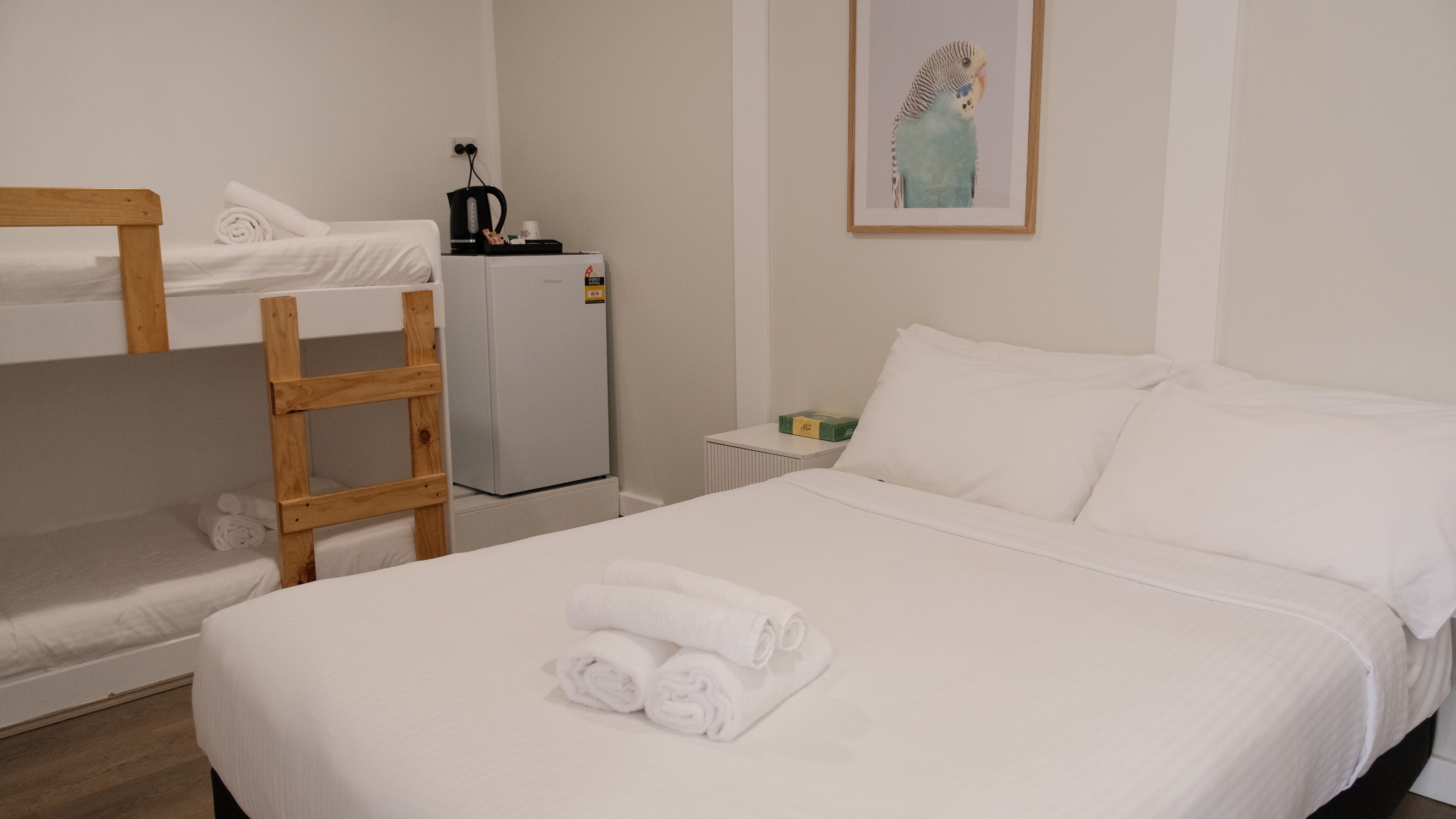 A hotel room with a single bed, a wooden chair, a small table with a kettle, and a refrigerator.