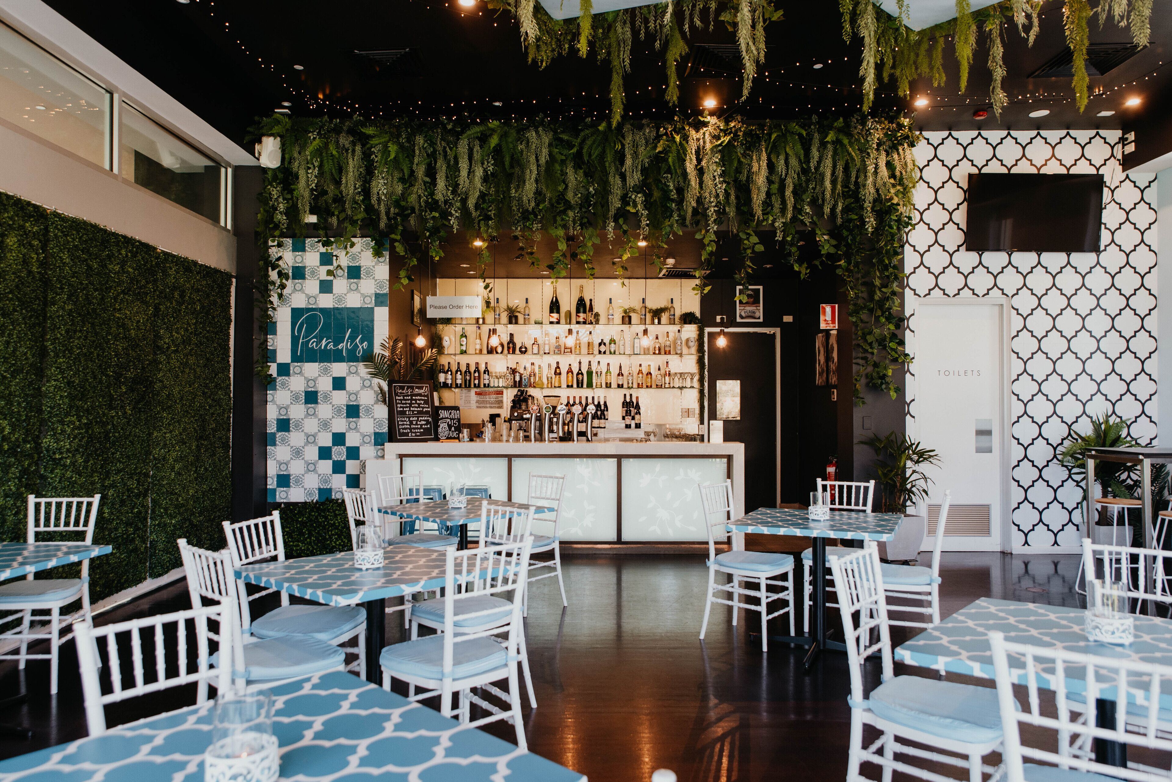 Un comedor interior con sillas y mesas blancas, una barra con una variedad de botellas, y una pared verde con plantas colgantes.