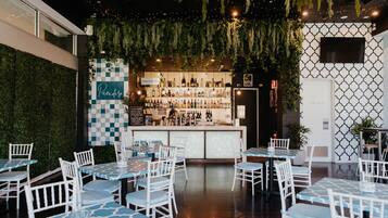 An indoor dining area with white chairs and tables, a bar with a variety of bottles, and a green wall with hanging plants.